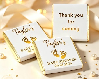 May include: Three square white chocolate favors with gold foil edges. Two feature the text Taylor's, baby footprints with heart cutouts, BABY SHOWER, and 06.01.2024. The third favor displays the gold text Thank you for coming in a centered layout.