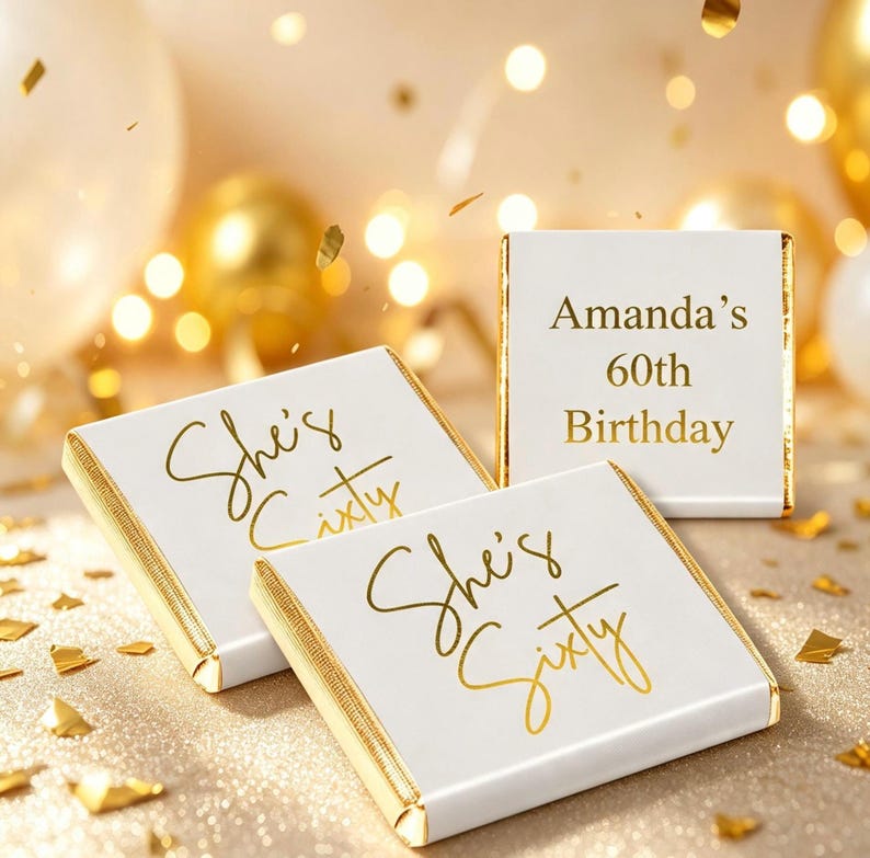 May include: Three square chocolate bars wrapped in white paper with gold foil edges. Two bars feature the gold script text She's Sixty, while the third bar displays the gold text Amanda's 60th Birthday in a clean, centered font style.