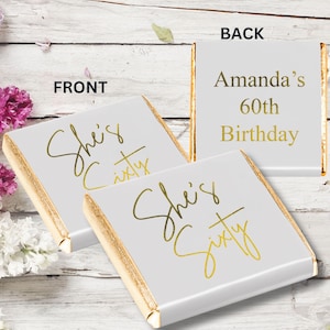 May include: Two square chocolate bars wrapped in white foil with gold metallic edges. The front wrappers feature the gold script text "She's Sixty". The back wrapper displays the gold text "Amanda's 60th Birthday" in a clean, sans-serif font.