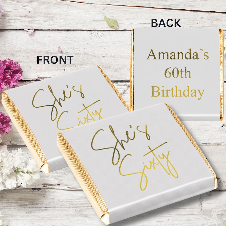 May include: Two square chocolate bars wrapped in white foil with gold metallic edges. The front wrappers feature the gold script text "She's Sixty". The back wrapper displays the gold text "Amanda's 60th Birthday" in a clean, sans-serif font.