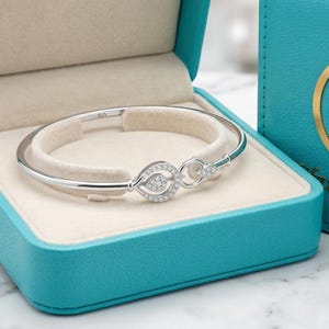 Könnte beinhalten: Ein silbernes Armband mit diamantartigem Akzent wird in einer offenen türkisfarbenen Schmuckschatulle mit cremefarbenem Innenraum präsentiert. Daneben befindet sich eine passende Schachtel mit goldenem Logo und dem Text "Hia GemsShop".