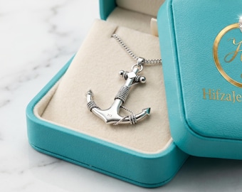 May include: A silver anchor pendant necklace displayed in a teal jewelry box. The anchor is detailed with wrapped wire and hangs from a delicate chain. The box is open, revealing a cream-colored interior. The box has the text "HifzaJewelsShop".