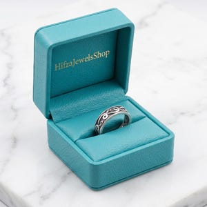Könnte beinhalten: Ein silberner Ring mit schwarzem Wirbelmuster in einer offenen, türkisfarbenen Ringschatulle. Die Schatulle hat ein türkisfarbenes Innenfutter und den goldenen Schriftzug "HifzaJewelsShop". Die Schatulle liegt auf einer Marmoroberfläche.