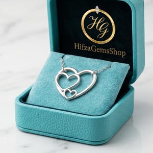 May include: A silver necklace with three heart pendants inside a teal jewelry box. The box is open, revealing a dark teal interior and the necklace resting on a blue velvet cushion. The box has the text "HifzaGemsShop".