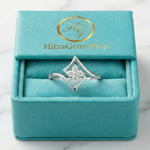 Könnte beinhalten: Ein silberner Ring mit einem einzigartigen geometrischen Design, der mit einer Ansammlung kleiner, funkelnder Steine in der Mitte versehen ist. Der Ring wird in einer offenen, türkisfarbenen Schmuckschatulle mit dem Text "HifzaGemsShop" in goldfarbenen Buchstaben auf dem Deckel präsentiert.