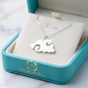 Può includere: Una collana color argento con un ciondolo a forma di nuvola con ritagli a spirale stilizzati, presentata in un portagioie color ottanio con il testo dorato HifzaJewelsShop stampato sul davanti. Il ciondolo pende da una sottile catenina argentata.