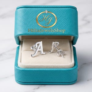 Könnte beinhalten: Ein Paar silberne Ohrstecker mit dem Buchstaben "A" in einer offenen türkisfarbenen Schmuckschatulle. Die Schatulle hat ein weißes Innenfutter und ein goldenes Logo mit der Aufschrift "PhizaJewelsShop". Die Ohrringe liegen auf einer weißen Oberfläche.