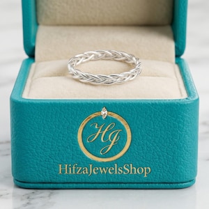 Könnte beinhalten: Ein silberner geflochtener Ring liegt in einer offenen türkisfarbenen Schmuckschatulle mit goldenem Logo und dem Text "HifzaJewelsShop". Der Ring ist mittig vor einem Marmorhintergrund platziert.