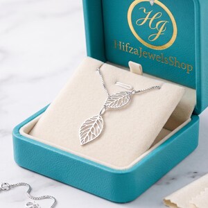 Könnte beinhalten: Eine silberfarbene Halskette mit zwei filigranen, durchbrochenen blattförmigen Anhängern. Die Kette liegt in einer türkisfarbenen Schmuckschatulle, auf deren Innendeckel der Text HifzaJewelsShop und ein Logo in Gold aufgedruckt sind.
