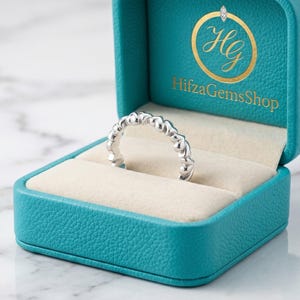 Könnte beinhalten: Ein silberner Ring mit sich wiederholendem herzförmigem Design wird in einer türkisfarbenen Schmuckschatulle präsentiert. Die Schatulle ist geöffnet und zeigt einen cremefarbenen Innenraum. Die Schatulle trägt den Text "HifzaGemsShop" und ein goldenes Logo.