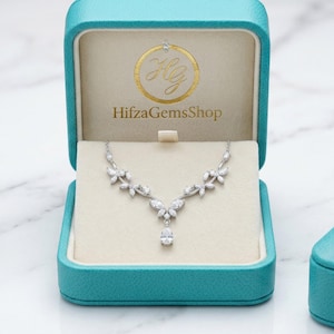 Könnte beinhalten: Eine silberne Halskette mit floralem Design und einem zentralen tropfenförmigen Anhänger wird in einer offenen, türkisfarbenen Schmuckschatulle präsentiert. Die Schatulle ist mit cremefarbenem Stoff ausgekleidet und trägt den Text "HifzaGemsShop". Daneben liegt eine passende geschlossene Schatulle.