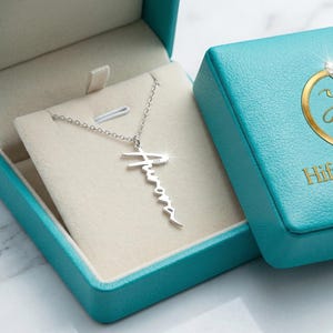 Könnte beinhalten: Eine silberfarbene Halskette mit Namensanhänger wird in einer offenen, türkisfarbenen Schmuckschatulle präsentiert. Die Schatulle hat eine hellfarbene Innenseite und einen passenden Deckel mit einem goldenen Logo mit der Aufschrift "HifzaGemsShop".