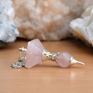 Pode incluir: Um pêndulo de quartzo rosa rosa com uma corrente prateada e uma parte superior adornada de prata e ouro. O pêndulo está sobre uma superfície de madeira.