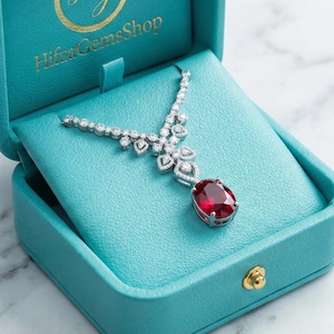 Könnte beinhalten: Eine silberne Halskette mit einem großen ovalen roten Edelsteinanhänger, präsentiert in einer offenen türkisfarbenen Schmuckschatulle. Die Halskette hat ein zartes Design mit kleinen, klaren Steinen. Die Schmuckschatulle hat einen goldenen Verschluss und den Text "HifaaGemsShop".