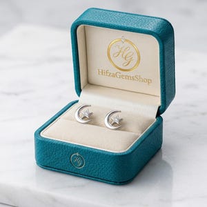 Könnte beinhalten: Silberne Ohrstecker mit Mond- und Sternmotiv, präsentiert in einer offenen, türkisfarbenen Schmuckschatulle mit cremefarbenem Innenraum. Die Schatulle zeigt das goldene Logo und den Shop-Namen "HifzaGemsShop".