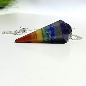 Könnte beinhalten: Ein sieben-Chakra-Kristallpendel mit einer silbernen Kette. Das Pendel ist ein spitzer Kegel mit Schichten aus roten, orangefarbenen, gelben, grünen, blauen, indigofarbenen und violetten Kristallen.