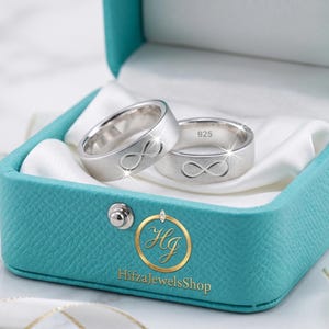 Peut inclure: Deux bagues en argent avec un symbole infini, présentées dans un écrin turquoise ouvert, doublé de satin blanc. L'écrin est posé sur une surface en marbre. Un ruban blanc avec des bordures dorées est disposé autour de la boîte.