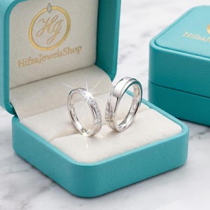 Könnte beinhalten: Zwei silberfarbene Ringe in einer türkisfarbenen Schmuckschatulle mit dem goldenen Logo HifraJewelsShop auf dem Innendeckel. Ein Ring hat ein schmales Band mit kleinen eingelassenen Steinen, der andere ist ein breiteres, glattes, poliertes Band.