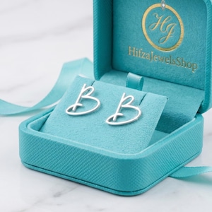 Könnte beinhalten: Silberfarbene Ohrringe in Form der Buchstaben "BB" in einer offenen, türkisfarbenen Schmuckschatulle präsentiert. Die Schatulle hat einen türkisfarbenen Samteinsatz, der die Ohrringe hält. Ein türkisfarbenes Band ist um die Schatulle gebunden, und das Logo "HifzaJewelsShop" ist sichtbar.