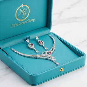 Könnte beinhalten: Ein silbernes Halsketten- und Ohrring-Set mit roten Edelsteinen, präsentiert in einer offenen türkisfarbenen Schmuckschatulle. Die Schatulle trägt den Text "Hifza GemsShop" in goldfarbenen Buchstaben. Der Schmuck liegt auf einem türkisfarbenen Hintergrund.