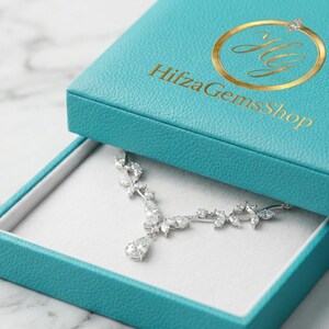 Könnte beinhalten: Geöffnete, türkisfarbene Schmuckschatulle mit einer silbernen Halskette. Auf dem Deckel der Schatulle befindet sich ein goldenes Logo und der Text "Hifza Gems Shop". Die Halskette hat ein florales Design mit klaren Edelsteinen.