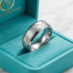 Puede incluir: Un anillo plateado con un diseño geométrico se muestra dentro de una caja turquesa. El anillo tiene un acabado de metal cepillado con una serie de líneas anguladas grabadas alrededor de la banda. La caja tiene un interior suave y aterciopelado.