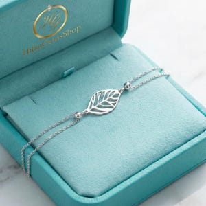 Könnte beinhalten: Ein silbernes Blattarmband in einer türkisfarbenen Schmuckschatulle. Das Armband hat einen filigranen Blattanhänger und kleine Kugelakzente. Die Schatulle ist geöffnet und zeigt ein weiches, türkisfarbenes Innere und den Markennamen "Hittac Gems Shop" in Gold.