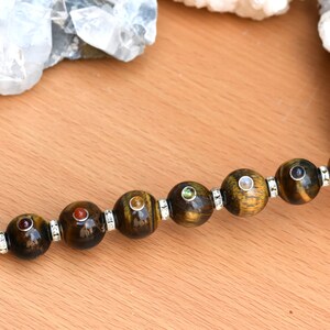 May include: A crystal wand with a clear quartz point and a silver crown. The wand is adorned with seven tiger's eye beads, each with a different colored gemstone inset.