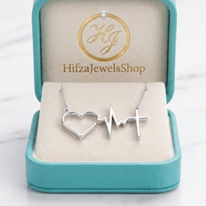 May include: A silver-toned necklace featuring a pendant with a heart, an EKG heartbeat line, and a cross, displayed inside a teal jewelry box. The interior of the box lid features a gold logo with the text HifzaJewelsShop printed below it.