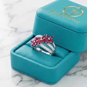 Könnte beinhalten: Ein silberner Ring mit einem floralen Design mit roten Edelsteinen und kleinen weißen Steinen, präsentiert in einer offenen türkisfarbenen Schmuckschatulle. Die Schatulle hat ein goldenes Logo auf dem Deckel und liegt auf einer Marmoroberfläche.