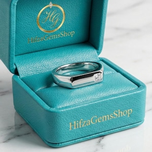 Könnte beinhalten: Ein silberner Ring mit rechteckigem Design und einem kleinen, klaren Edelstein wird in einer offenen, türkisfarbenen Schmuckschatulle präsentiert. Auf der Schatulle steht der Text "HifzaGemsShop". Der Ring liegt auf einem türkisfarbenen Innenraum.