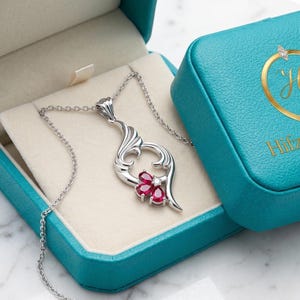 Könnte beinhalten: Eine silberne Anhänger-Halskette mit floralem Design und roten Edelsteinen, präsentiert in einer offenen türkisfarbenen Schmuckschatulle. Die Schatulle steht auf einer Marmoroberfläche, mit dem Markennamen "HifzaGemsShop".