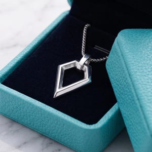 Könnte beinhalten: Ein silberfarbener Anhänger in geometrischer Drachenform an einer passenden Kette, präsentiert in einer strukturierten türkisfarbenen Schmuckschatulle mit schwarzem Samtinnenfutter. Der Deckel zeigt ein goldenes Logo mit dem Text HifzaJew.