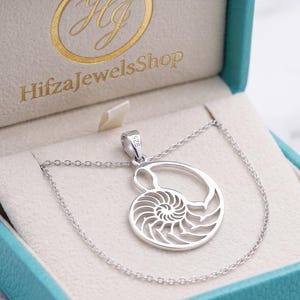 Könnte beinhalten: Eine silberne Nautilus-Muschel-Halskette mit einem detaillierten spiralförmigen Ausschnitt-Design. Der Anhänger ist mit 925 gestempelt und liegt in einer türkisfarbenen Schmuckschatulle mit dem goldenen Schriftzug HifzaJewelsShop.
