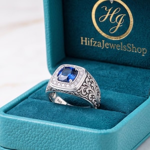 Könnte beinhalten: Ein Silberring mit einem blauen Edelstein im Kissenschliff, umgeben von kleinen weißen Steinen, mit filigranen Rankengravuren auf der Ringschiene und einem 925-Stempel, präsentiert in einer türkisfarbenen Schmuckschatulle mit goldenem Schriftzug HifzaJewelsShop.