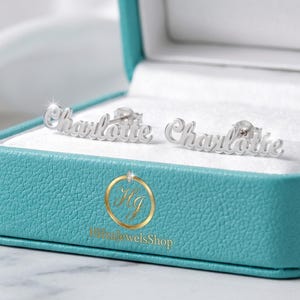 Puede incluir: Un par de pendientes plateados con el nombre "Charlotte" escrito en cursiva, presentados en una caja de joyería turquesa con interior blanco. Los pendientes se exhiben sobre una superficie blanca.