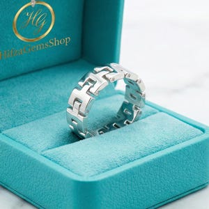 Könnte beinhalten: Ein silberfarbener Ring mit geometrischem Design liegt in einer türkisfarbenen Schmuckschatulle. Der Ring hat ein sich wiederholendes Muster aus ineinandergreifenden Formen. Die Schatulle hat ein weiches, samtiges Innenfutter und den Text "HilzaGemsShop".