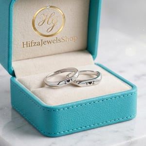 Könnte beinhalten: Zwei silberne Ringe mit den Buchstaben "Y" und "Z" und einem Herzsymbol liegen in einer offenen, türkisfarbenen Schmuckschatulle. Die Schatulle hat ein cremefarbenes Innenfutter. Die Ringe liegen auf einer weißen Oberfläche.