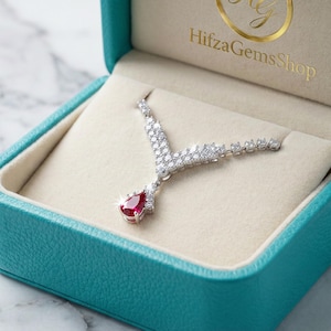 Könnte beinhalten: Eine silberne Halskette mit einem tropfenförmigen Rubinanhänger liegt in einer türkisfarbenen Schmuckschatulle. Die Halskette hat ein V-förmiges Design mit zahlreichen kleinen, klaren Steinen. Die Schatulle ist mit cremefarbenem Stoff ausgekleidet.