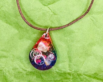 Puede incluir: Collar con colgante en forma de lágrima y cordón marrón. El colgante presenta una colorida mezcla de rojo, naranja, azul y morado, con cristales incrustados y un diseño de espiral plateada. El fondo es verde texturizado.