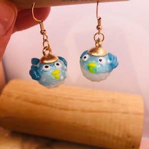 May include: A pair of handmade earrings featuring small, round, blue and white bird-like figures with gold-colored caps. The earrings have gold-colored hooks and are displayed against a light background.