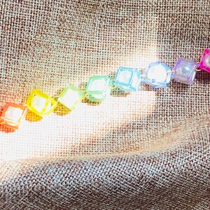 Peut inclure: Un bracelet composé d'une série de perles carrées translucides et multicolores disposées en dégradé arc-en-ciel, contenant chacune une perle sphérique blanche. Les perles sont reliées par une chaîne en métal doré avec un fermoir mousqueton.