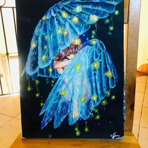 Op de afbeelding: Een tekening van een persoon met blauwe engelen vleugels, versierd met gele lichten, tegen een zwarte achtergrond. De vleugels zijn gedetailleerd met doorschijnende veren. Het kunstwerk staat op een spiraalgebonden schetsboek, op een houten oppervlak.