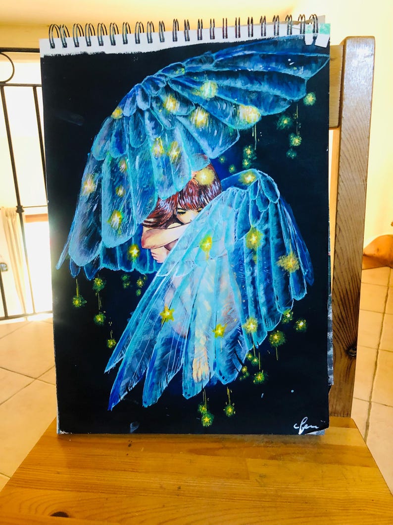 Op de afbeelding: Een tekening van een persoon met blauwe engelen vleugels, versierd met gele lichten, tegen een zwarte achtergrond. De vleugels zijn gedetailleerd met doorschijnende veren. Het kunstwerk staat op een spiraalgebonden schetsboek, op een houten oppervlak.