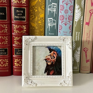 Op de afbeelding: Een wit fotolijstje met een decoratieve rand en een klein schilderij van twee mensen onder een paraplu. Het lijstje staat op een plank met een rij boeken erachter.