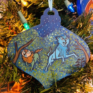 May include: A blue and green wooden Christmas ornament with a painted scene of two wolves in a forest. The wolves are painted in a stylized, folk art style. The ornament is decorated with white snowflakes and yellow stars.