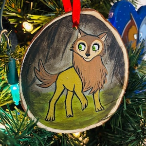 May include: A hand-painted wooden ornament featuring a yellow wolf with brown fur and green eyes. The wolf is standing in a dark forest setting with green grass and brown trees.