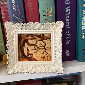 May include: A white framed picture with a whimsical illustration of two figures standing under a bare tree branch. The illustration is in a muted color palette of browns, yellows, and greens. The frame has a distressed white finish and ornate detailing.