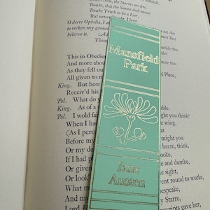 May include: A teal blue bookmark with gold lettering that reads "Mansfield Park" and "Jane Austen". The bookmark features a floral design with a gold outline.