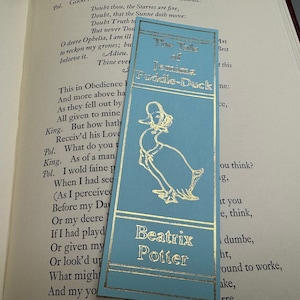May include: A blue bookmark with gold lettering featuring a cartoon illustration of a duck wearing a bonnet. The bookmark has the title "The Tale of Jemima Puddle-Duck" and the author's name "Beatrix Potter".